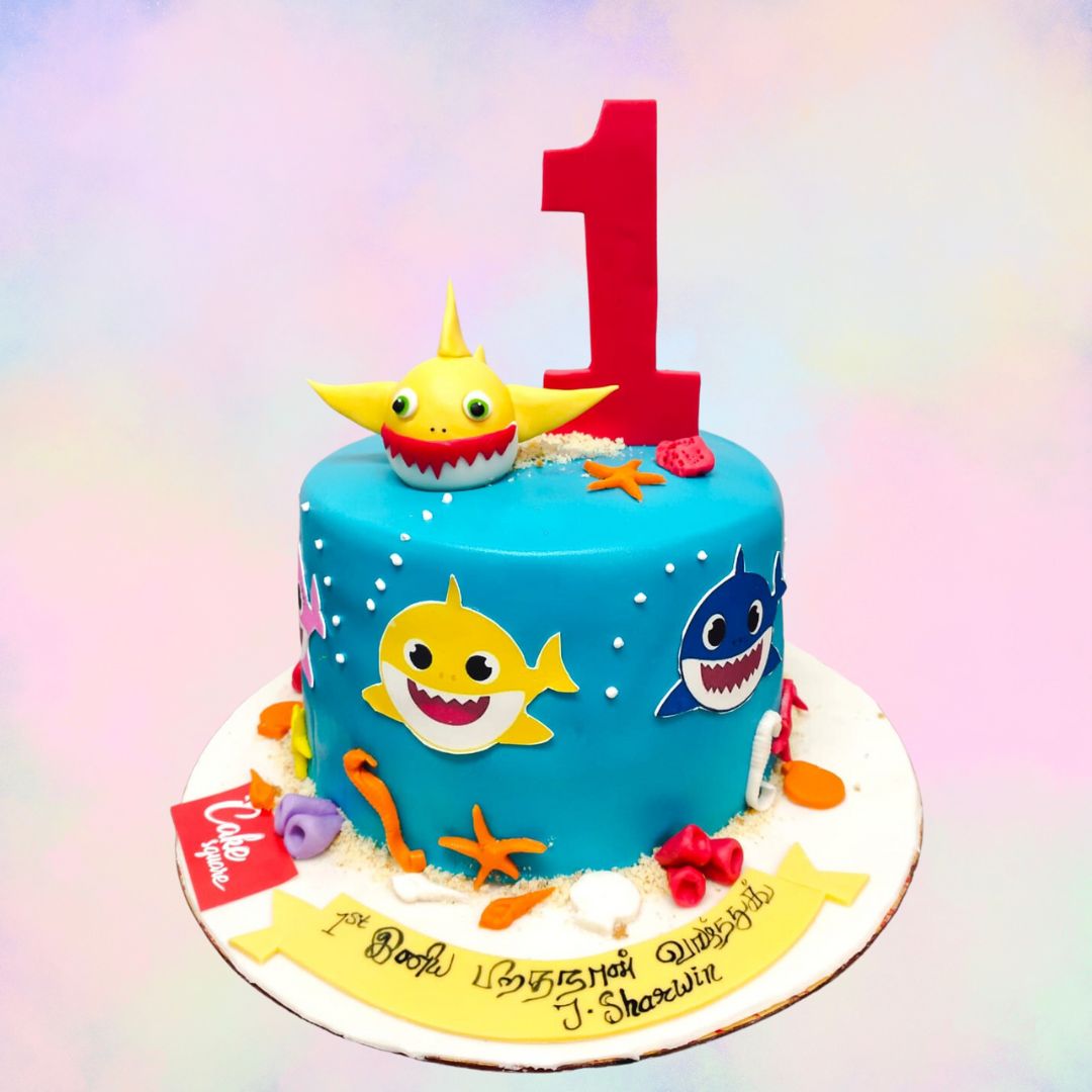 1 Kg Baby Shark Theme Kids First Birthday Cake, Adorable blue and white Baby Shark-themed cake with smiling shark characters, ocean waves, and a "1" topper, perfect for a child's first birthday celebration.