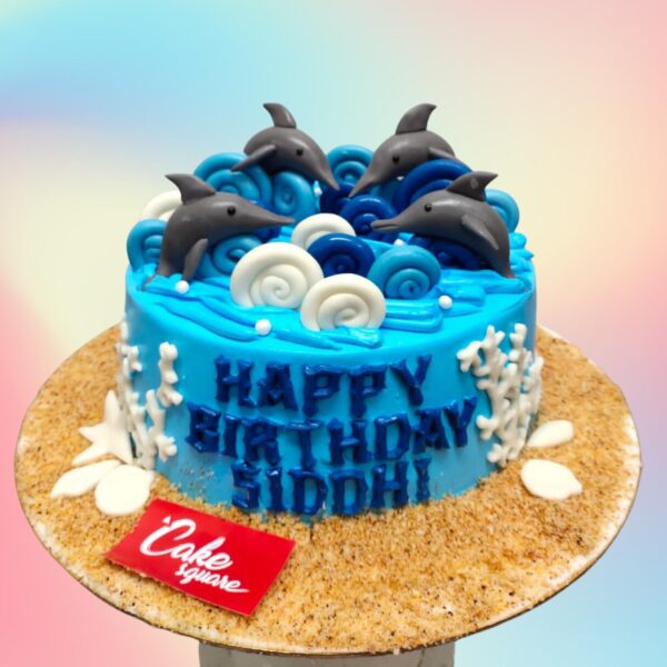 1 Kg Dolphin Theme Birthday Cake with a 3D fondant dolphin leaping from waves of blue buttercream. Decorated with edible seashells, starfish, and coral. "Happy Birthday" written on a fondant beach with a sandy texture.