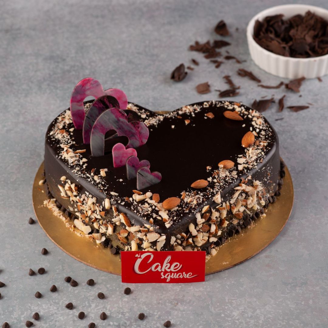 A large heart-shaped 1 Kg Chocolate Birthday Cake with smooth frosting, chocolate curl decorations, and crushed almonds all around.