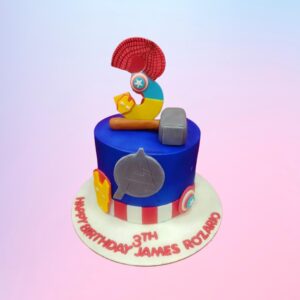 Perfect Avengers Theme Boys Birthday Cake 1 Kg. Features edible images of Captain America's shield, Thor's hammer, and Iron Man's arc reactor. Blue fondant base with red and gold accents. "Happy Birthday" written in comic-style font.