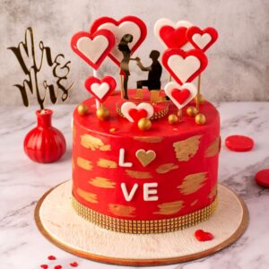 A red cake filled with hearts and a proposing topper on a beautiful butterscotch Gold Love 1 Kg Anniversary Cake by Cake Square team.