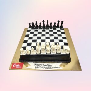 Spectacular Chess Board Birthday Cake 2 Kg with alternating black and white fondant squares. Edible chess pieces are arranged on top, with a golden "Happy Birthday" plaque placed diagonally across the board.