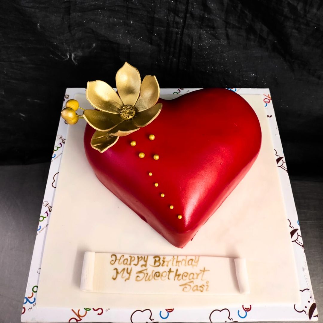 A red heart adorned with a big golden flower and droplets of gold sprinkles on top is Style 1Kg Anniversary Cake made by Cake Square team.