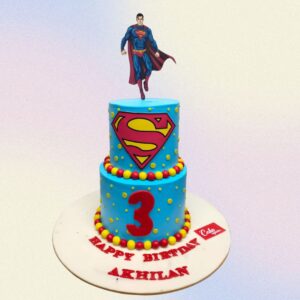 Stylish Super man Theme Birthday Cake 3 Kg. A rectangular cake decorated to resemble Superman's iconic costume. The top is covered in blue fondant with a raised red fondant "S" shield. The sides feature a red cape effect. Edible cityscapes and "Happy Birthday" text complete the heroic design.