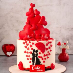 Kissing couple under an unbrella with red hearts all over a white cake is Umbrella Love 1 Kg Anniversary Cake, made by Cake Square team.