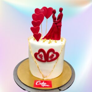 A white cake with two hearts in the front topped by fondant couple in red and adorned with hearts around them is Red Couple 1 Kg Anniversary Cakes by Cake Square team.