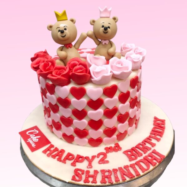 A single cake weighing 1 kg, designed with a teddy bear theme for twin girls' first birthday. The tier is pale pink, decorated with fondant layered cutouts. Two identical fondant teddy bears, wearing pink bows, sit atop the cake. Between them is a large fondant number "1" in pink. Fondant flowers and butterflies in shades of pink and white are scattered across . A banner reading "Happy 1st Birthday Twins" in pink lettering wraps around the bottom tier. Edible glitter adds a sparkly touch throughout.