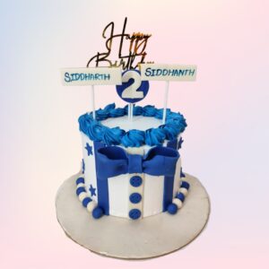 A round 1 kg cake designed for twin boys' second birthday. The cake is covered in light blue fondant. Two fondant teddy bears, each wearing a different colored bowtie, sit on top. Between them is a large fondant number "2" in white. The sides of the cake are decorated with fondant stars and moons in various shades of blue. Small toy cars and blocks are arranged around the base of the cake. The cake's border is piped with white buttercream in a scalloped pattern. A banner reading "Happy 2nd Birthday" in blue lettering stretches across the front of the cake.