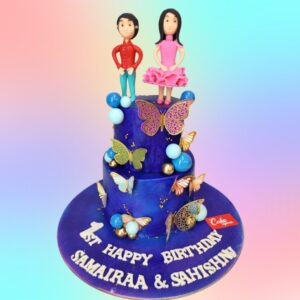 A one-tiered cake weighing 2 kg, designed for twins under 10. The bottom tier is blue, the top tier is pink, representing boy and girl twins. Both tiers are decorated with colorful fondant balloons, stars, and confetti. Two fondant child figures, one boy and one girl, stand on top. Between them is a large fondant number showing their age, in rainbow colors. The sides of the cake feature could be with fondant cut-outs of typical kids' interests - books, sports equipment, musical notes, and art supplies. A banner reading "Happy Birthday Twins" in multicolored letters wraps around the bottom tier.